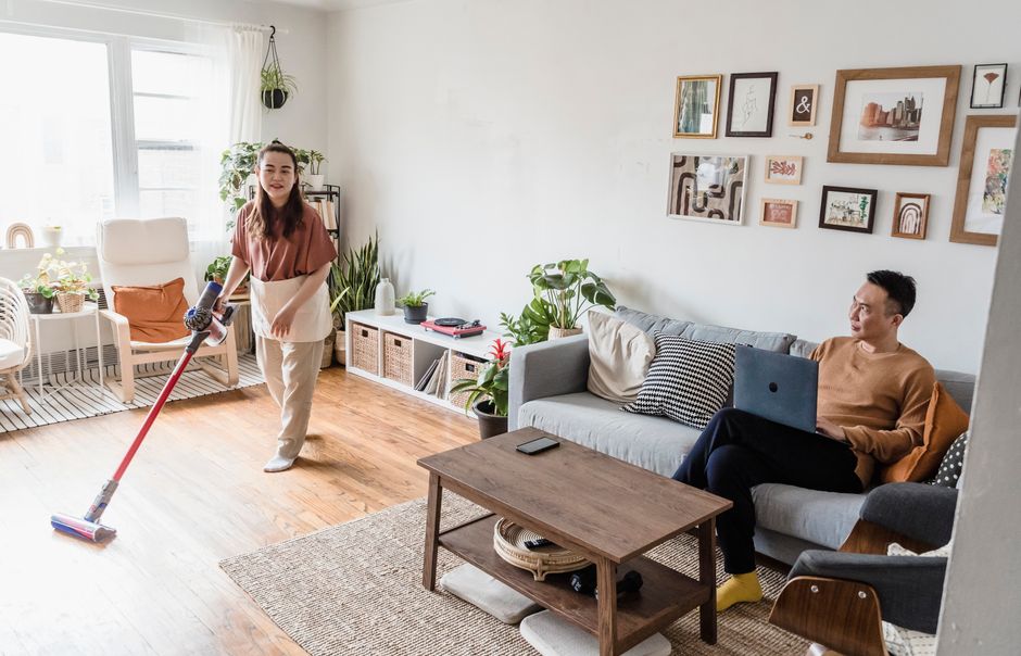 A man and woman indoors, one cleaning, the other using a laptop, in a modern living room.
